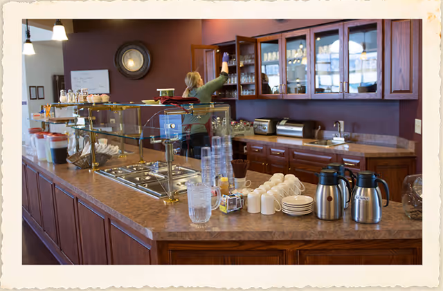 Cafe-style service counter with coffee urns, cups, pastries, and glass display in a senior living kitchen.
