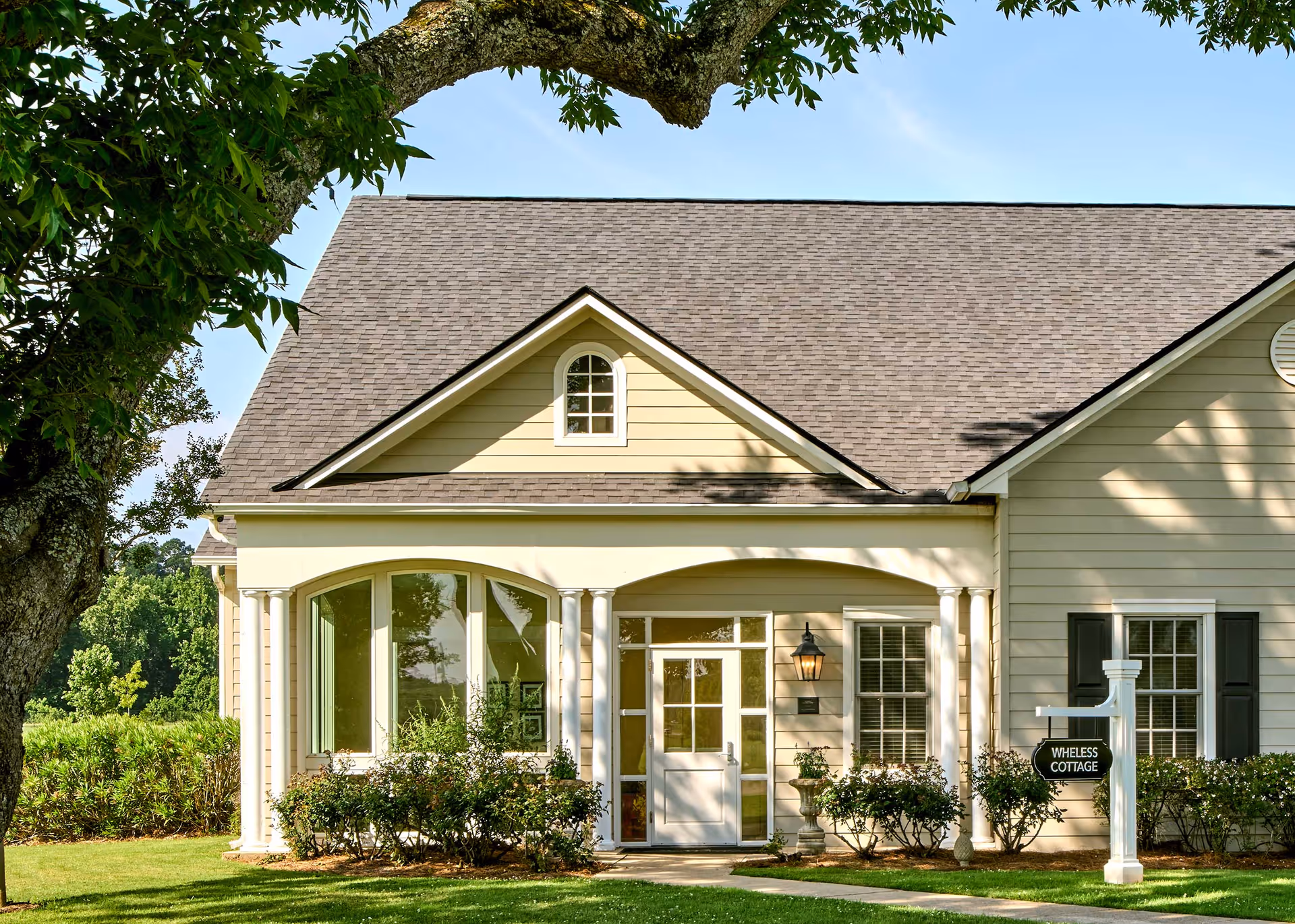 Front exterior of a beige cottage-style building with columns, arched windows, landscaping, and a 'WHELESS COTTAGE' sign.