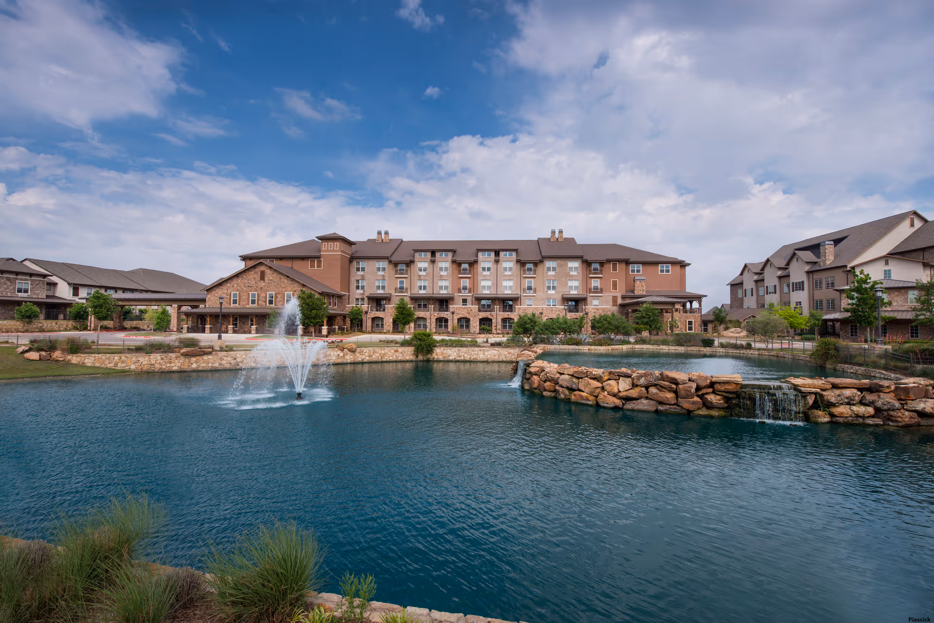 A multi-story senior living building fronting a pond with a central fountain and stone waterfall under a partly cloudy sky.