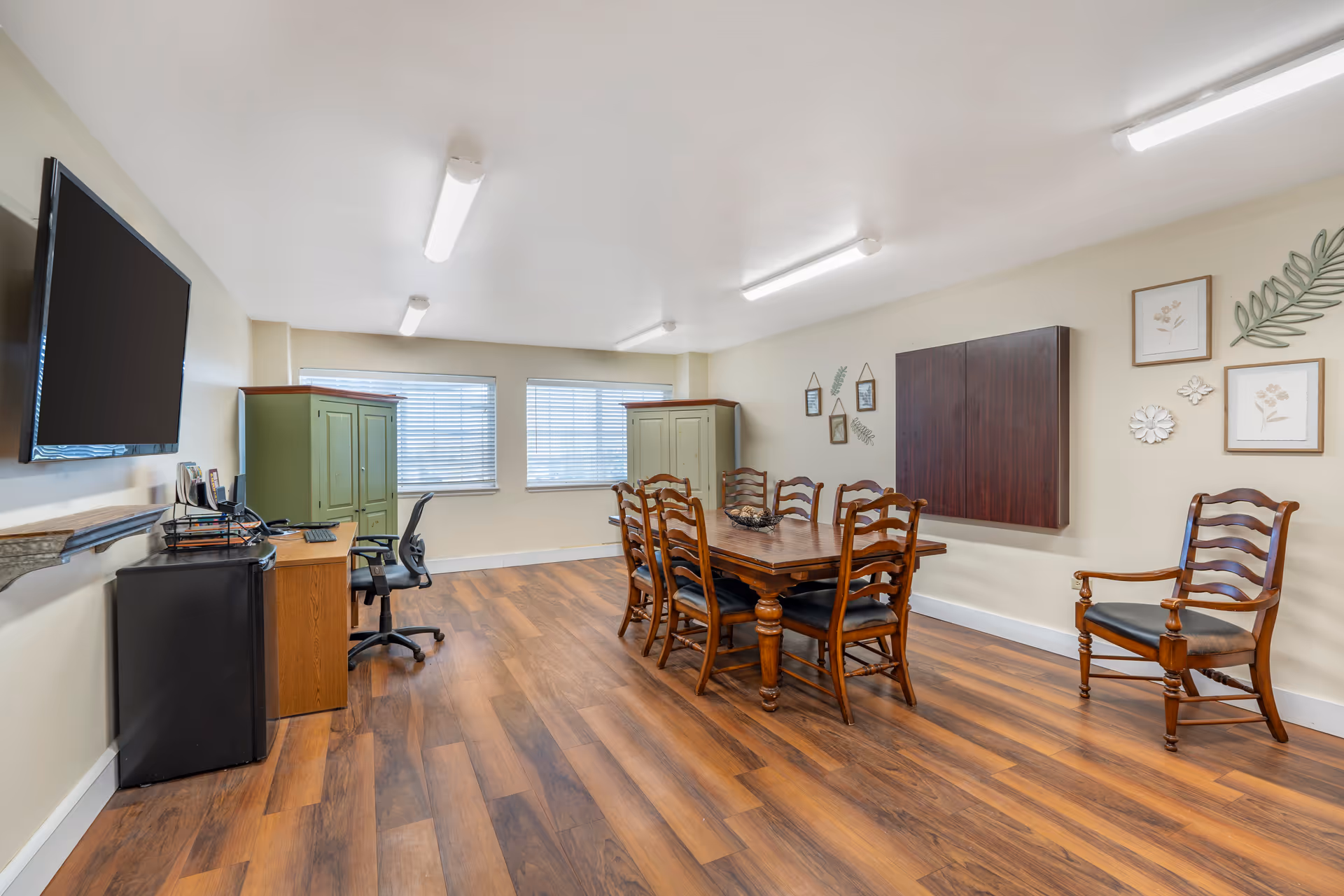 A well-lit room with wooden flooring featuring a wooden dining table surrounded by six chairs with dark cushions. There is an additional wooden chair with armrests to the right. On the left side, there is a desk with a computer chair, a small black refrigerator, and a wall-mounted flat-screen TV. Two green cabinets are positioned against the far wall beneath two windows with blinds. The walls are decorated with framed botanical prints and metal leaf accents.