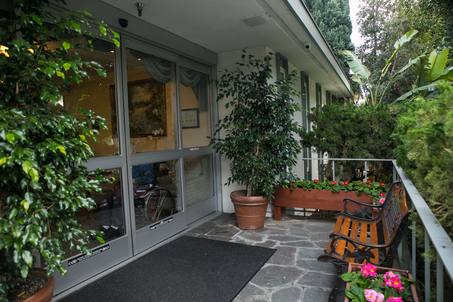 Entrance area of Brentwood Health Care Center showing glass doors leading inside. There are potted plants and flower boxes with colorful flowers along the stone-paved walkway. A wooden bench with decorative metal armrests is placed near the railing, surrounded by greenery.