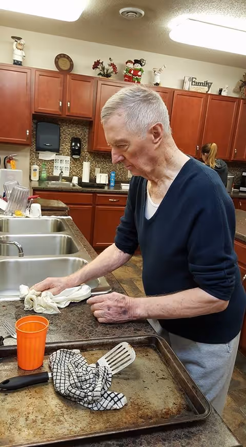An older man wipes a countertop in a communal kitchen with sinks, cabinets, and baking trays.