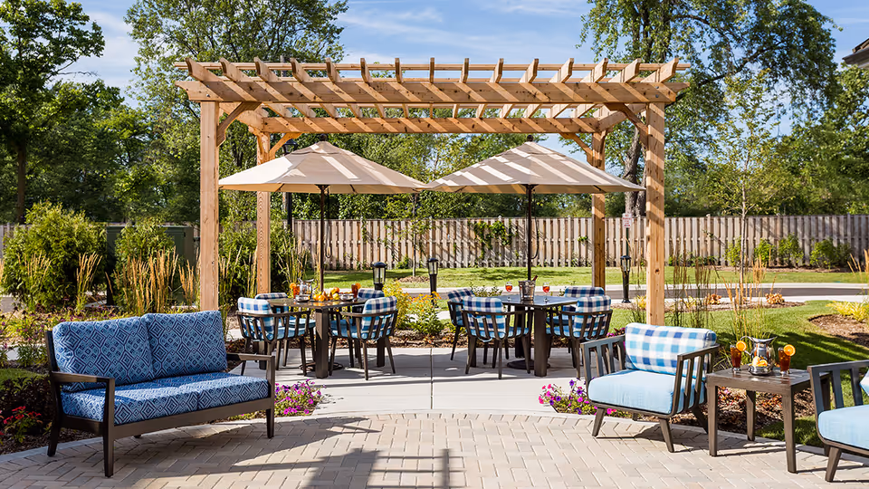 Outdoor patio area with a wooden pergola, two tables with umbrellas, and several chairs with blue patterned cushions. There are drinks and snacks on the tables, surrounded by greenery and a wooden fence in the background.