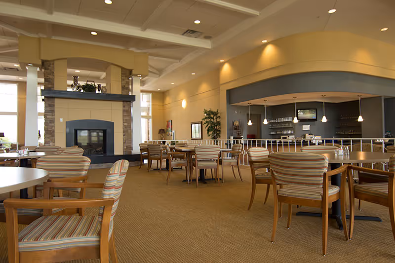Spacious dining room with tables, striped chairs, a fireplace and a bar area in a senior living community.