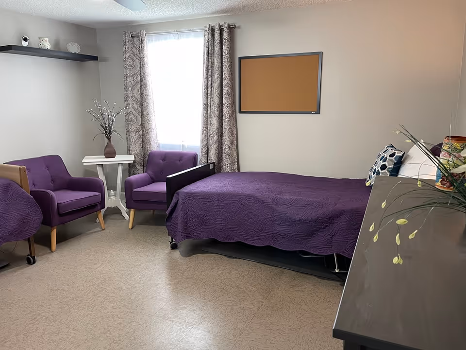 A bedroom in a senior facility with purple bedspread, chairs, and window curtains.