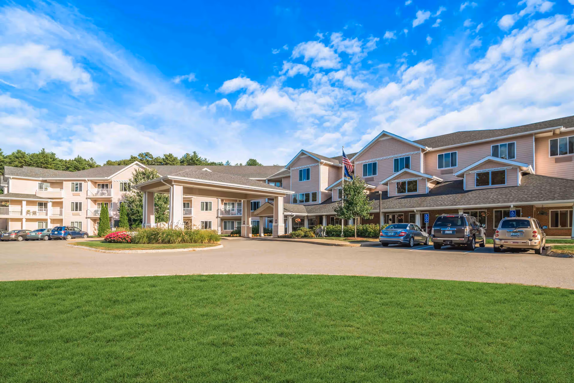 Front exterior of a senior living building with a covered entrance, flagpole, parked cars, and a grassy lawn under a blue sky.