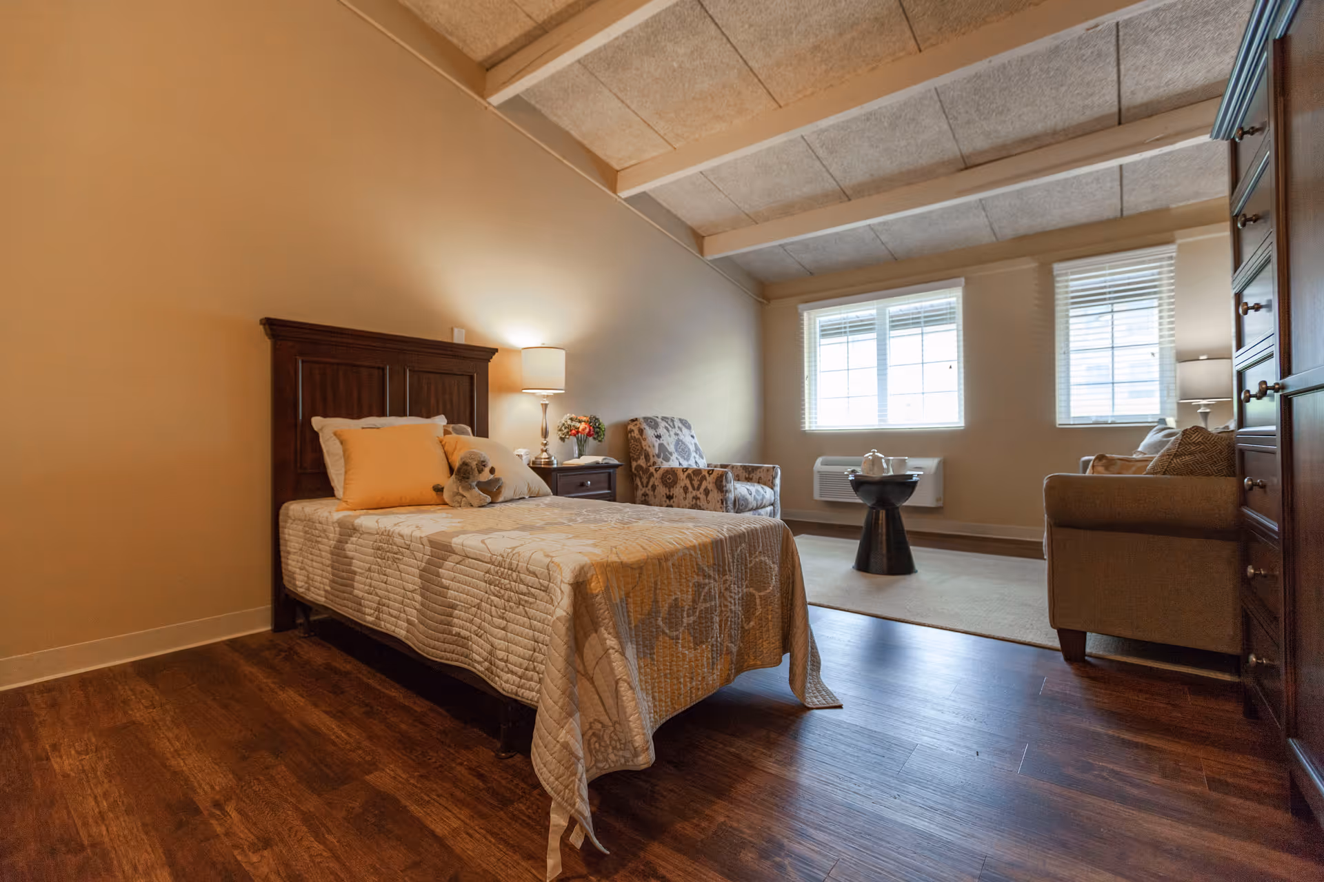 A furnished bedroom suite with a single bed, nightstand and lamp, a seating area with chairs and a sofa, and windows letting in daylight.