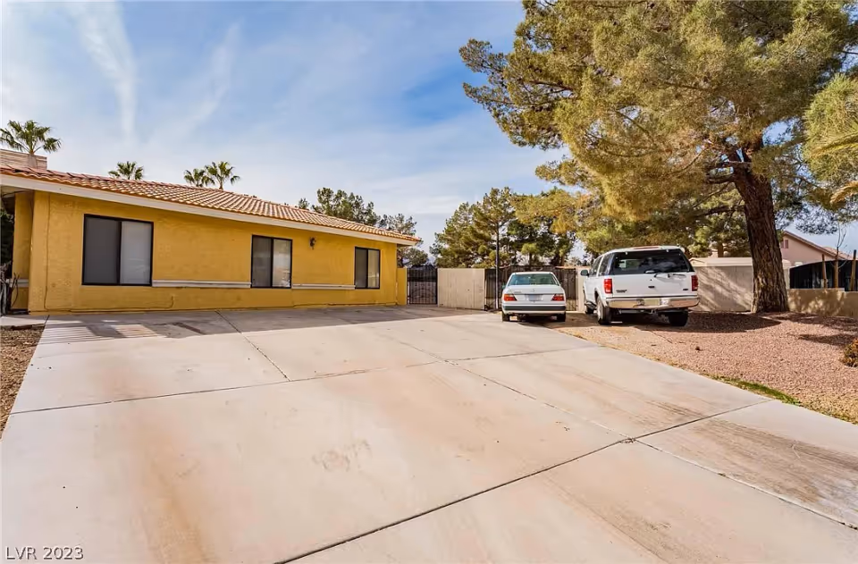 Single-story yellow stucco building with a large concrete driveway, two parked vehicles, and trees in the yard.