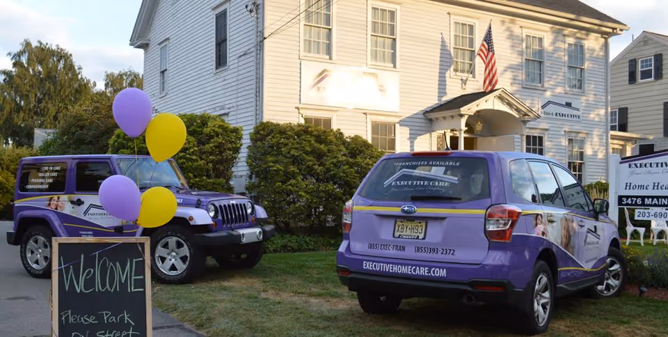 Two purple promotional vehicles parked along a white two-story building with balloons and a chalkboard sign.