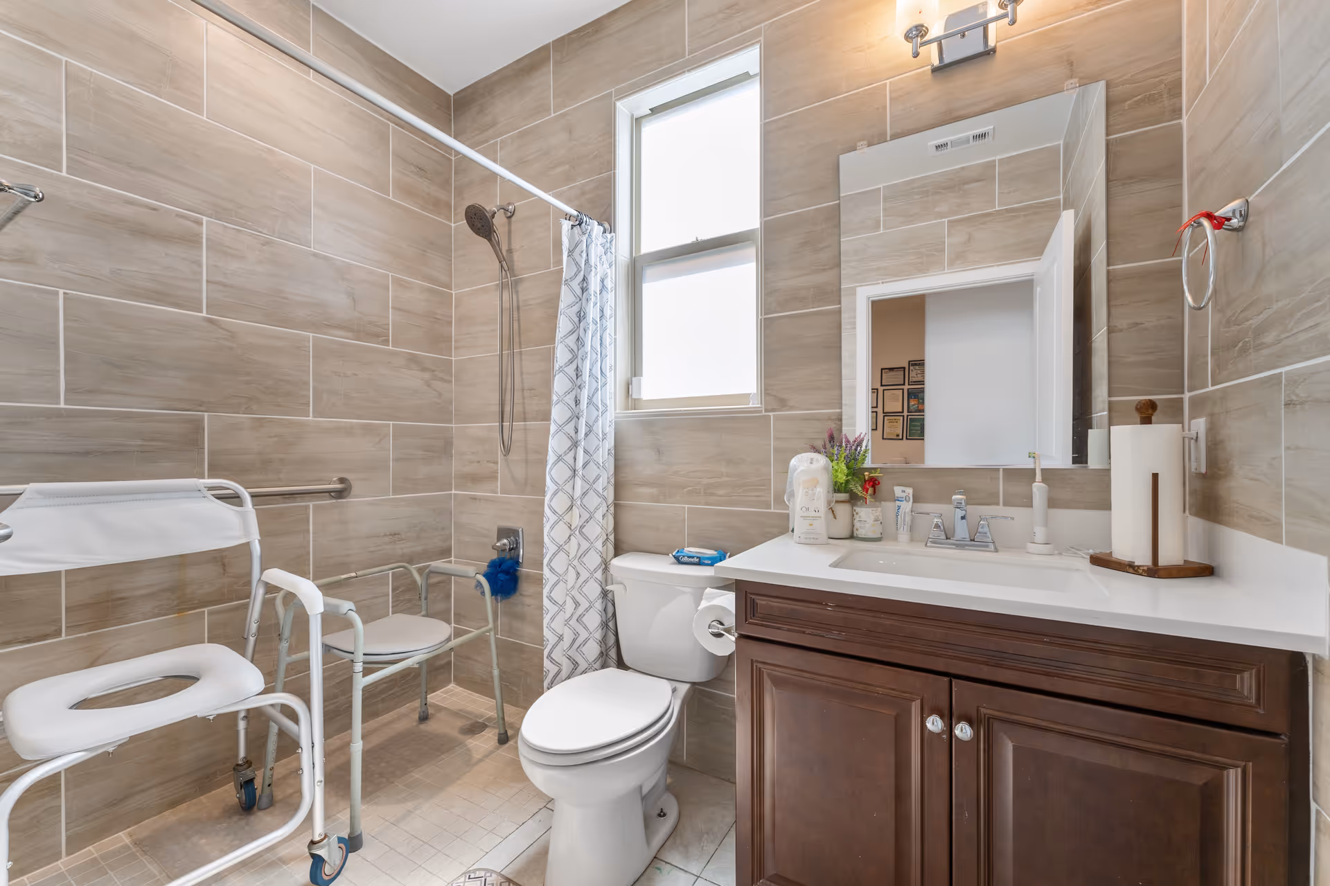 Accessible bathroom with walker, shower chair, toilet, and vanity in beige tiled room.