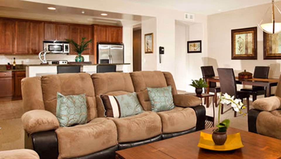 Open-concept living area with a beige reclining sofa, coffee table, dining table, and a kitchen with stainless steel appliances in the background.