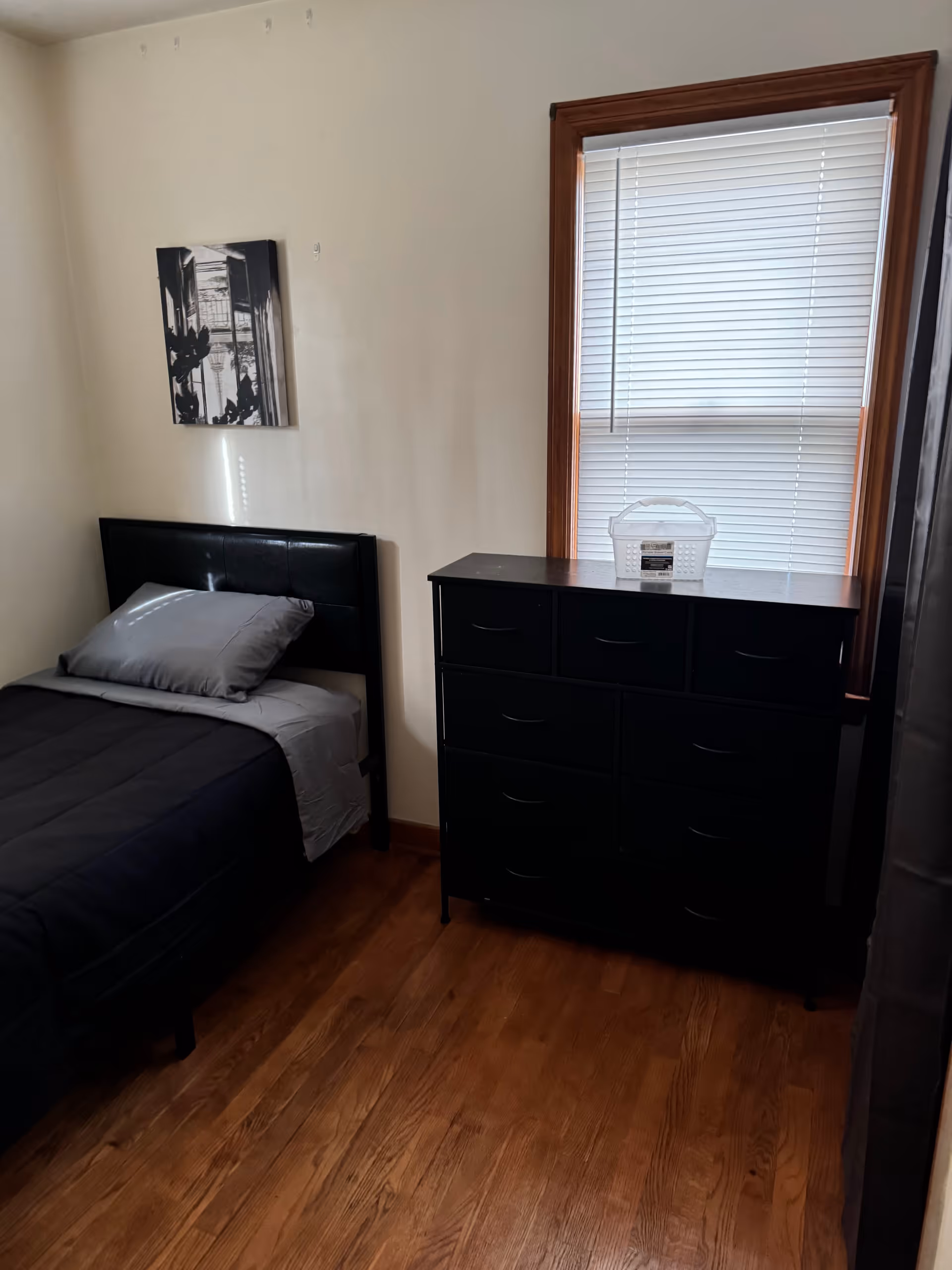 Small bedroom with a twin bed, black dresser, wooden floor, and window with blinds.