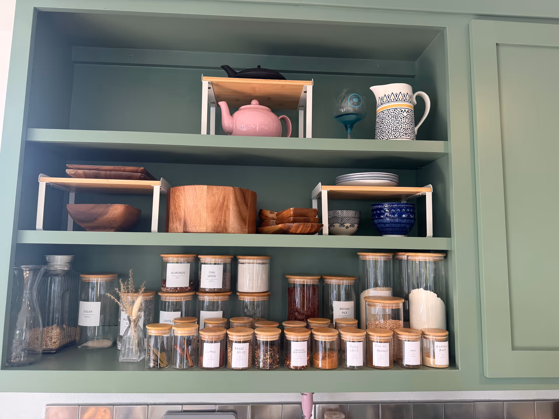 Green open kitchen cabinet with shelves holding dishes, jars, and a pink teapot.