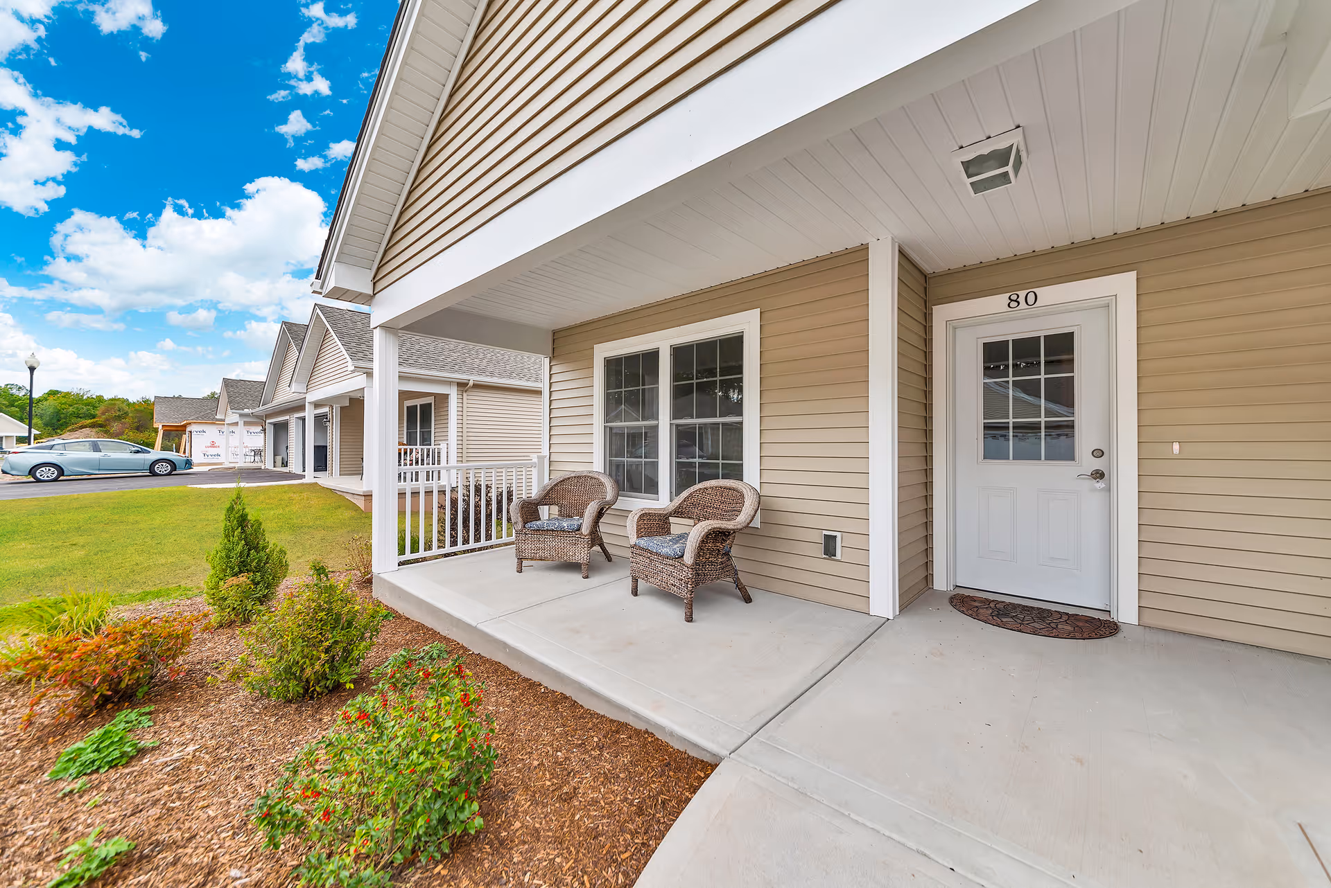Front porch of a residential unit at Gardens of Wilbraham with two wicker chairs, a white door labeled 80, beige siding, and a small garden area with shrubs and flowers. A car and other similar units are visible in the background under a partly cloudy blue sky.