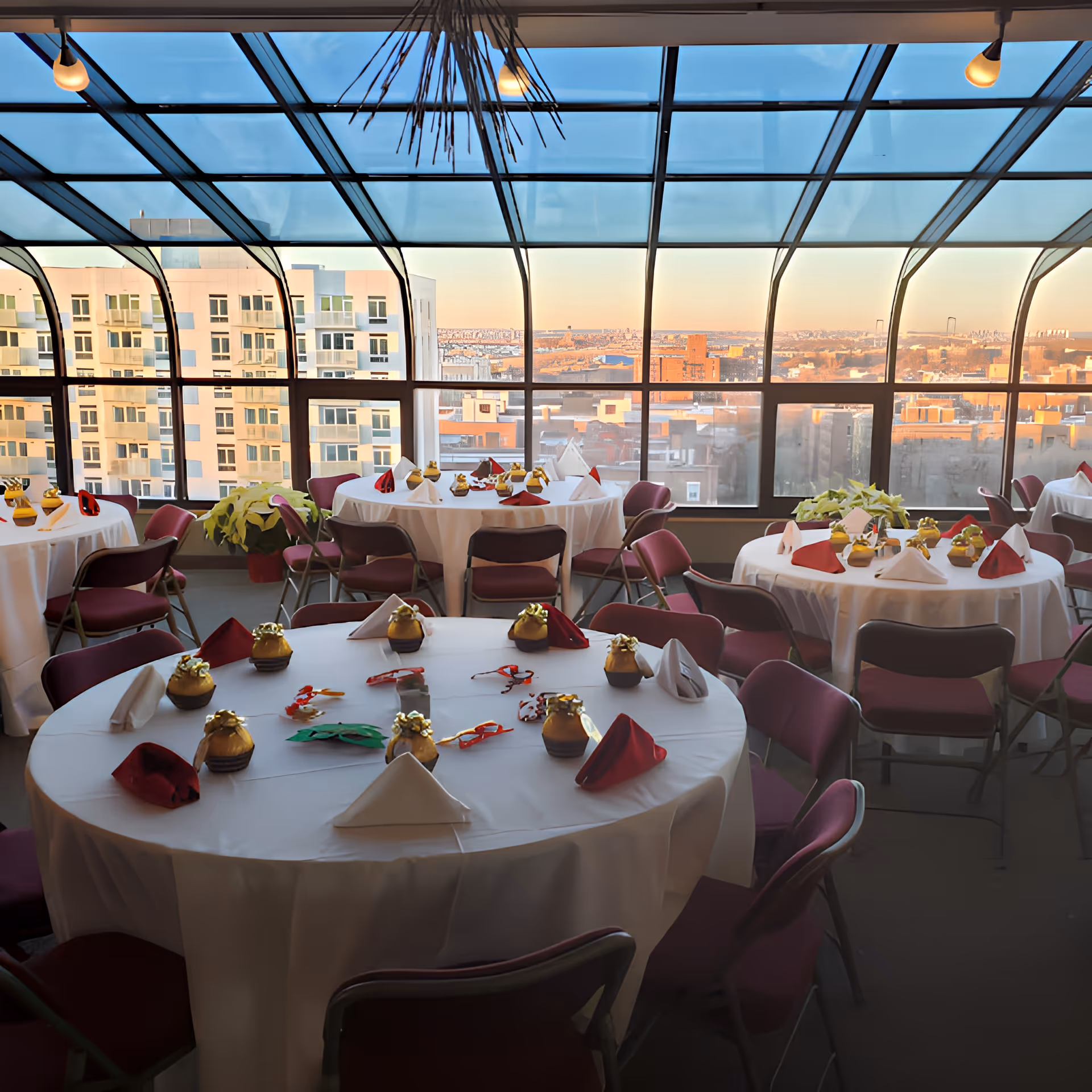 Sunlit dining room with round tables set with white tablecloths, folded napkins and small centerpieces beneath a large curved glass skylight overlooking city buildings.