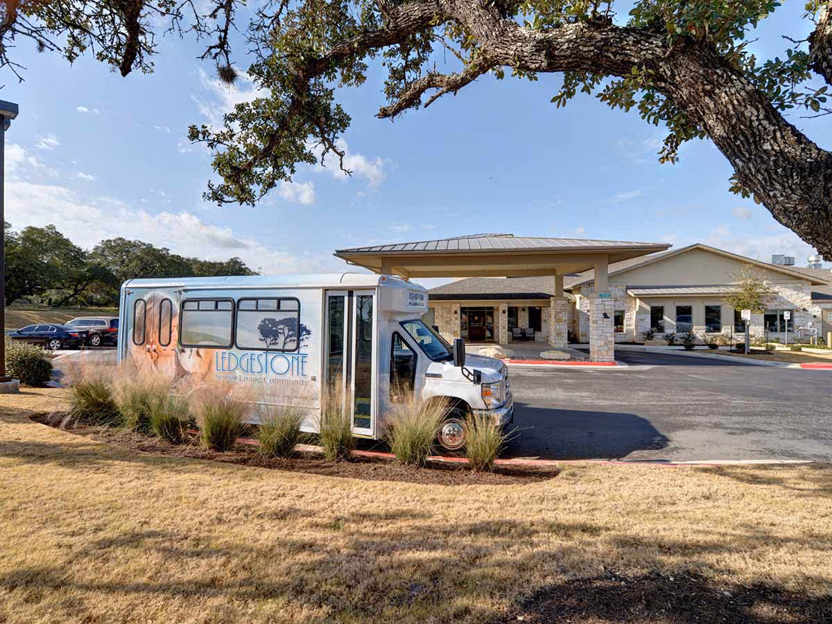 Shuttle van parked outside the covered entrance of the Ledgestone Senior Living building with trees and lawn.