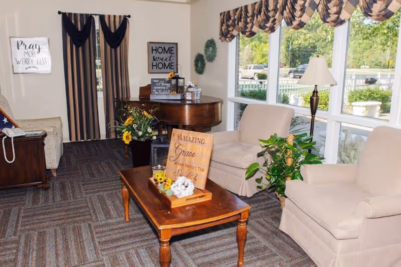 Cozy senior living lounge with beige seating, coffee table, piano, and large windows.