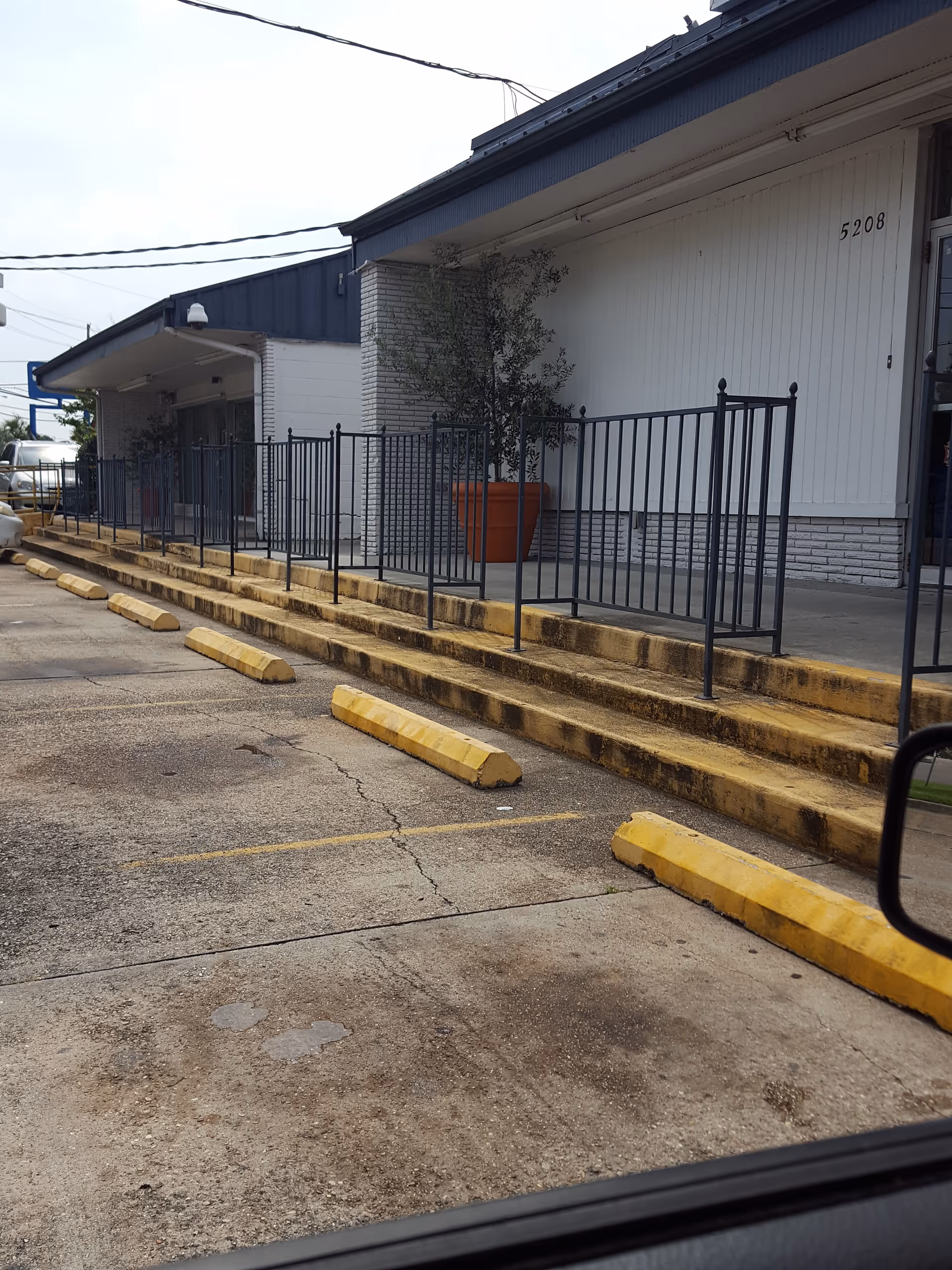 Front exterior of a senior facility with steps, railing, and a large potted plant.