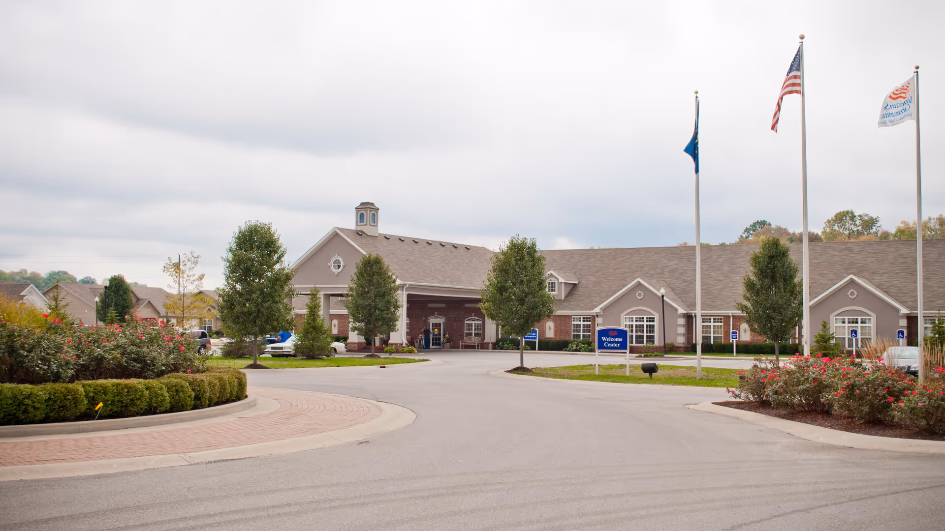 Meadow Lakes senior living facility exterior with entrance driveway, landscaping, and flags.