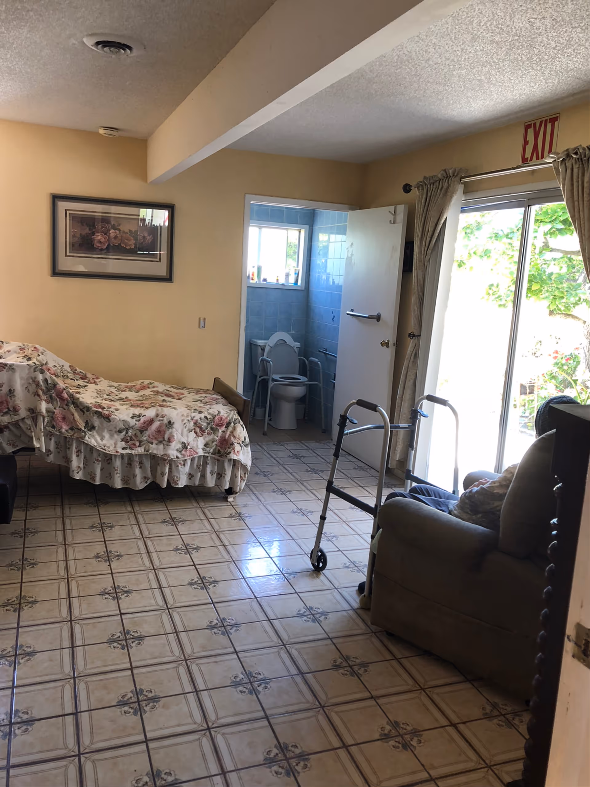 Furnished senior living room with floral bed, walker, sofa, and open bathroom door.