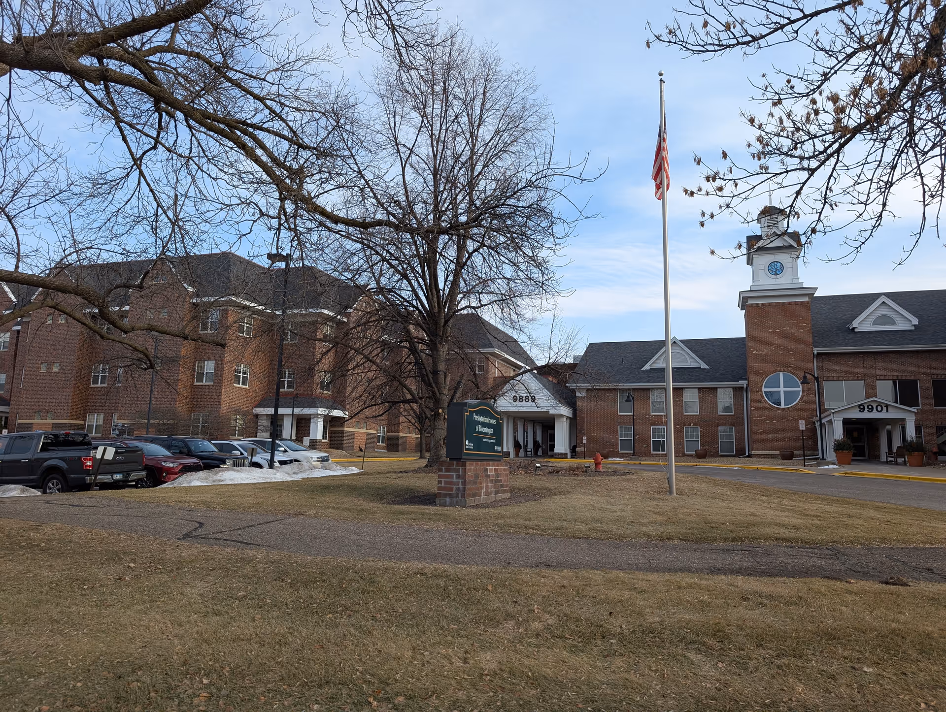 Brick senior living facility building and entrance with a flagpole, a sign on the lawn, parked cars, and leafless trees.