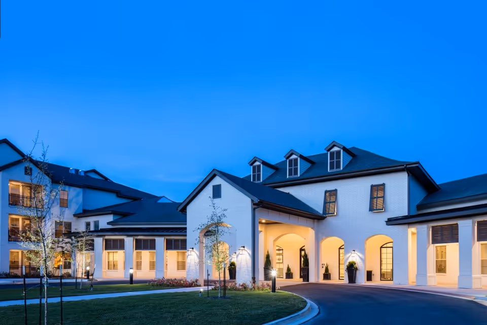 Well-lit front entrance of a multi-story senior living building at dusk with arched entryways and landscaped grounds.