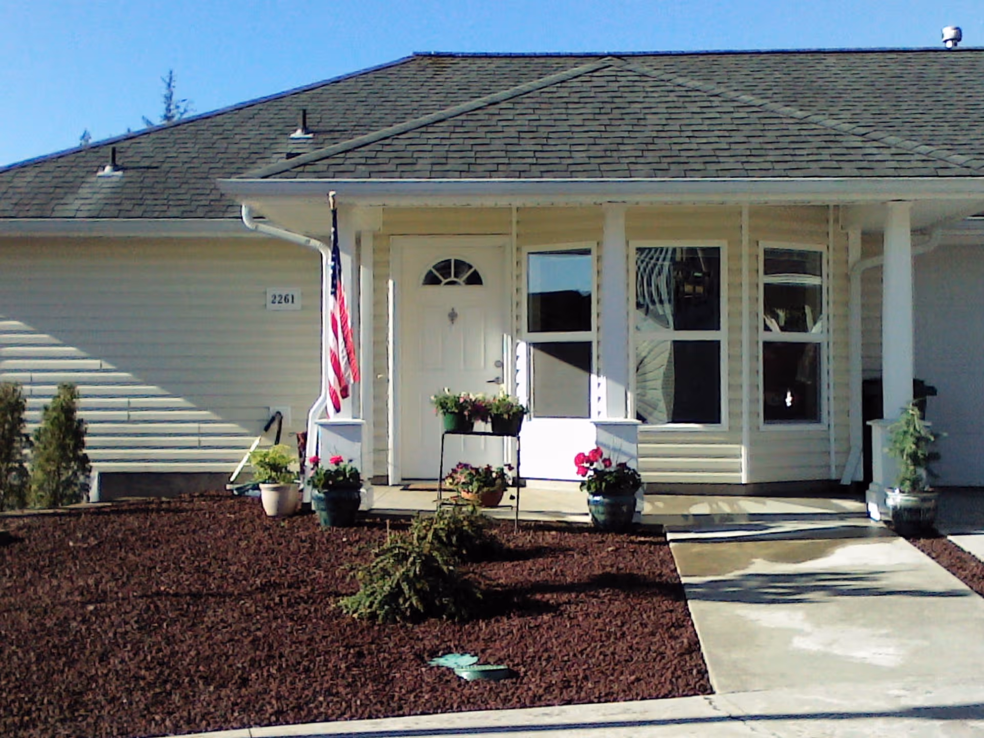 Front exterior of a single-story retirement community with a porch, flag, and potted flowers.