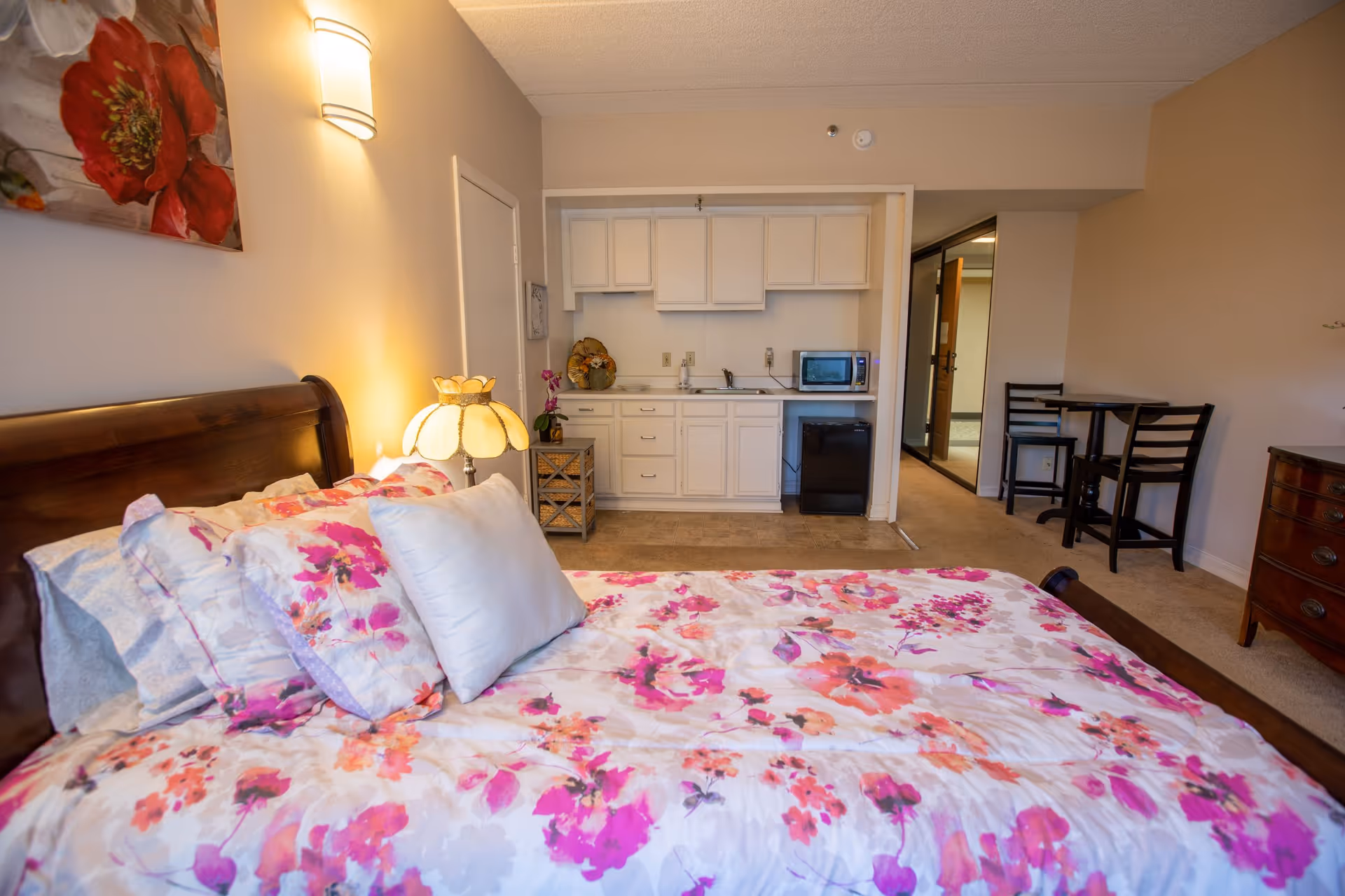 Senior living studio bedroom with floral bedspread, bedside lamp, and kitchenette.
