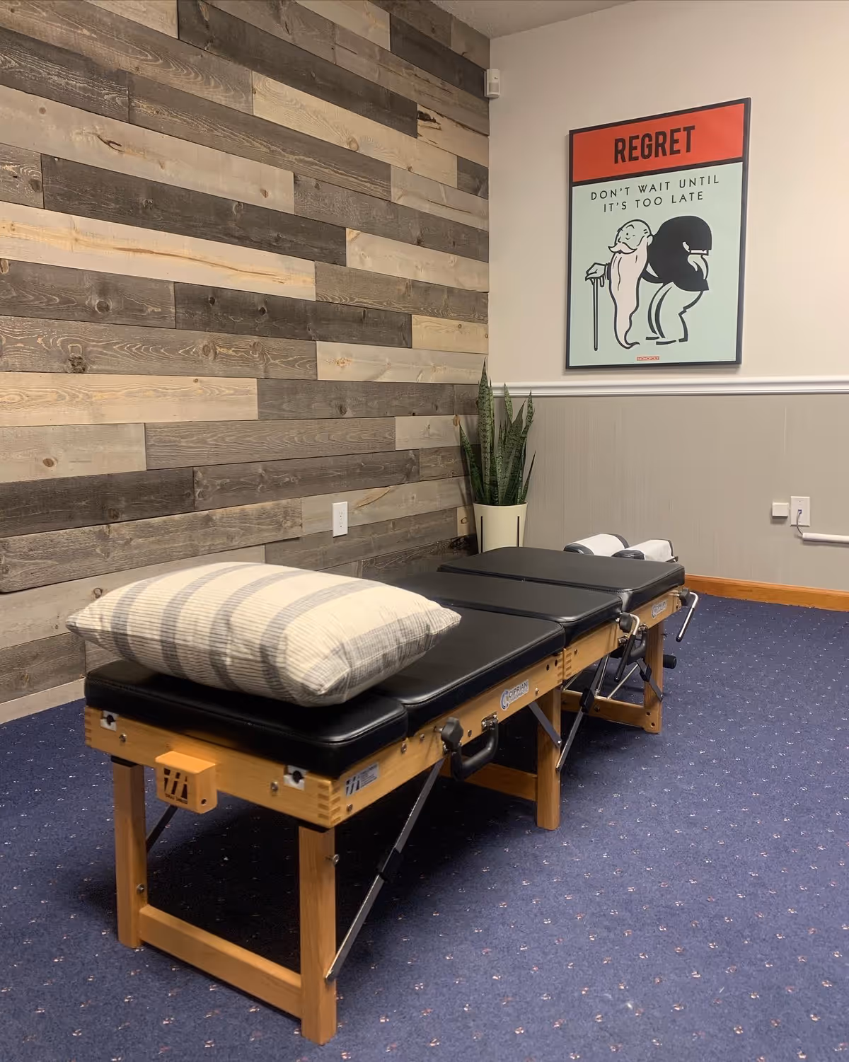 Wood-paneled wall, therapy table with pillow, plant, and a 'Regret' poster in a quiet room.