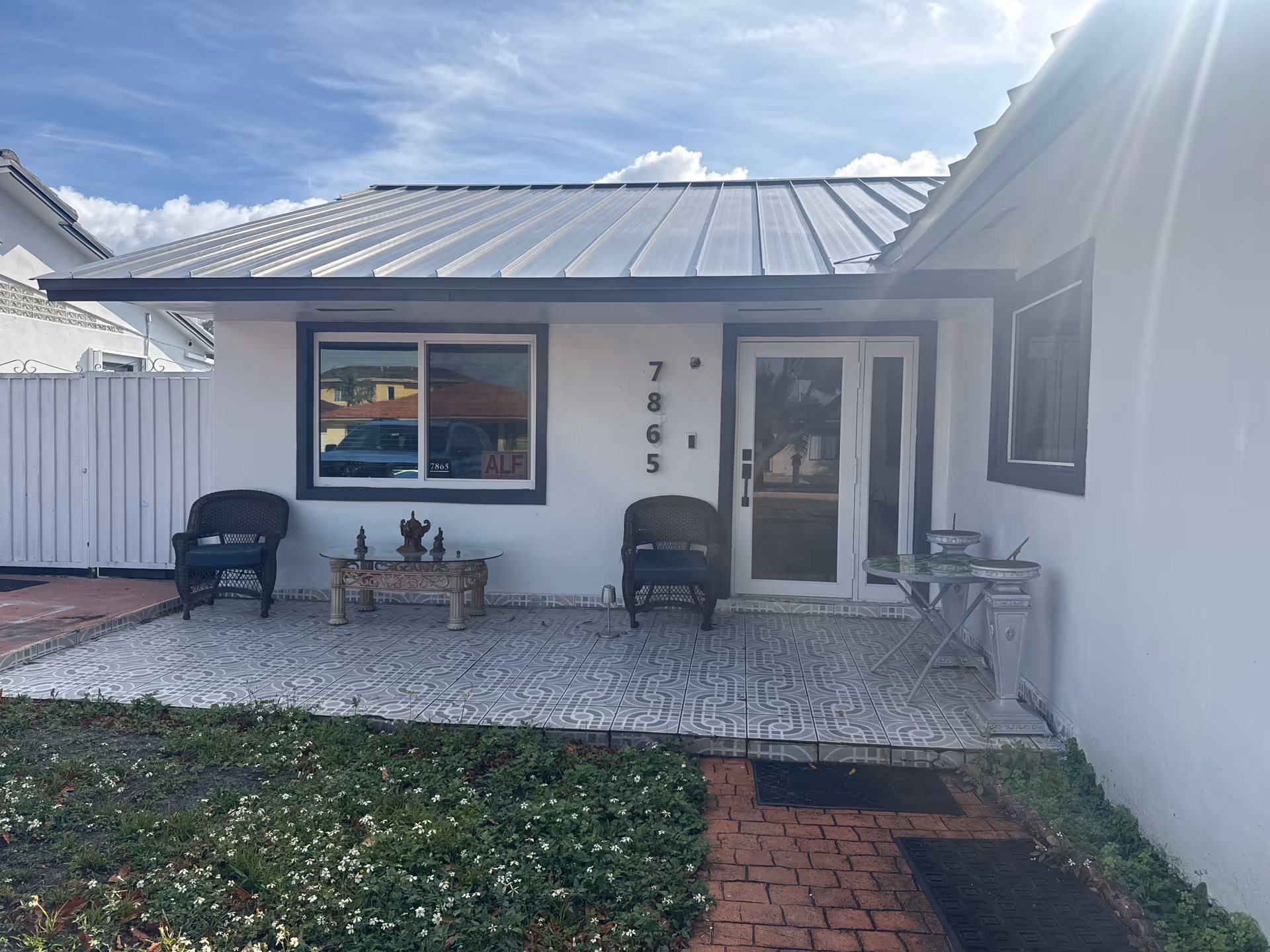 White single-story building with a covered porch, two chairs, a small table, and a glass door.