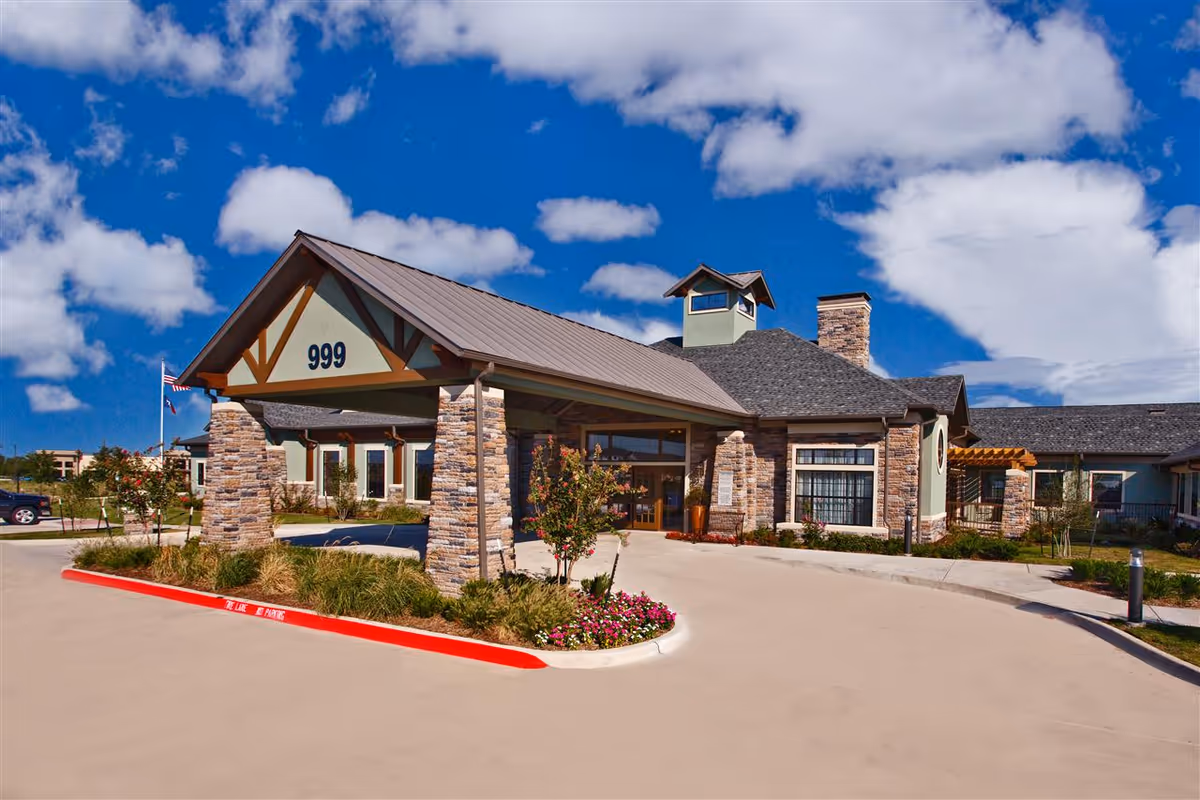 Exterior view of a senior living facility building with stone pillars and a covered entrance marked with the number 999. The building has a pitched roof and a chimney, with landscaped greenery and flowers around the driveway under a partly cloudy blue sky.