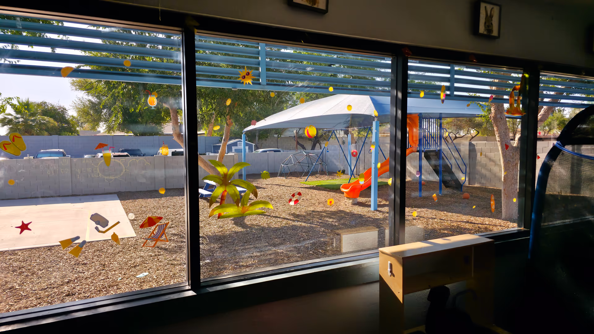 View through large windows shows a sunny outdoor playground with slides and canopy.