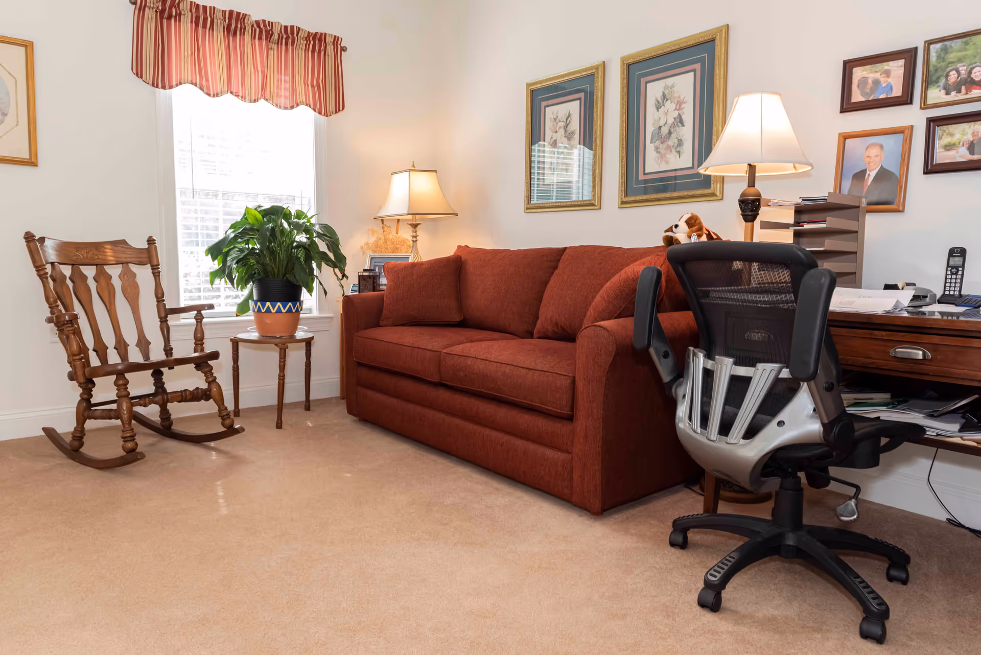 A cozy room with a red sofa, a wooden rocking chair, a small table with a potted plant, a desk with an office chair, a lamp, and framed pictures on the wall.