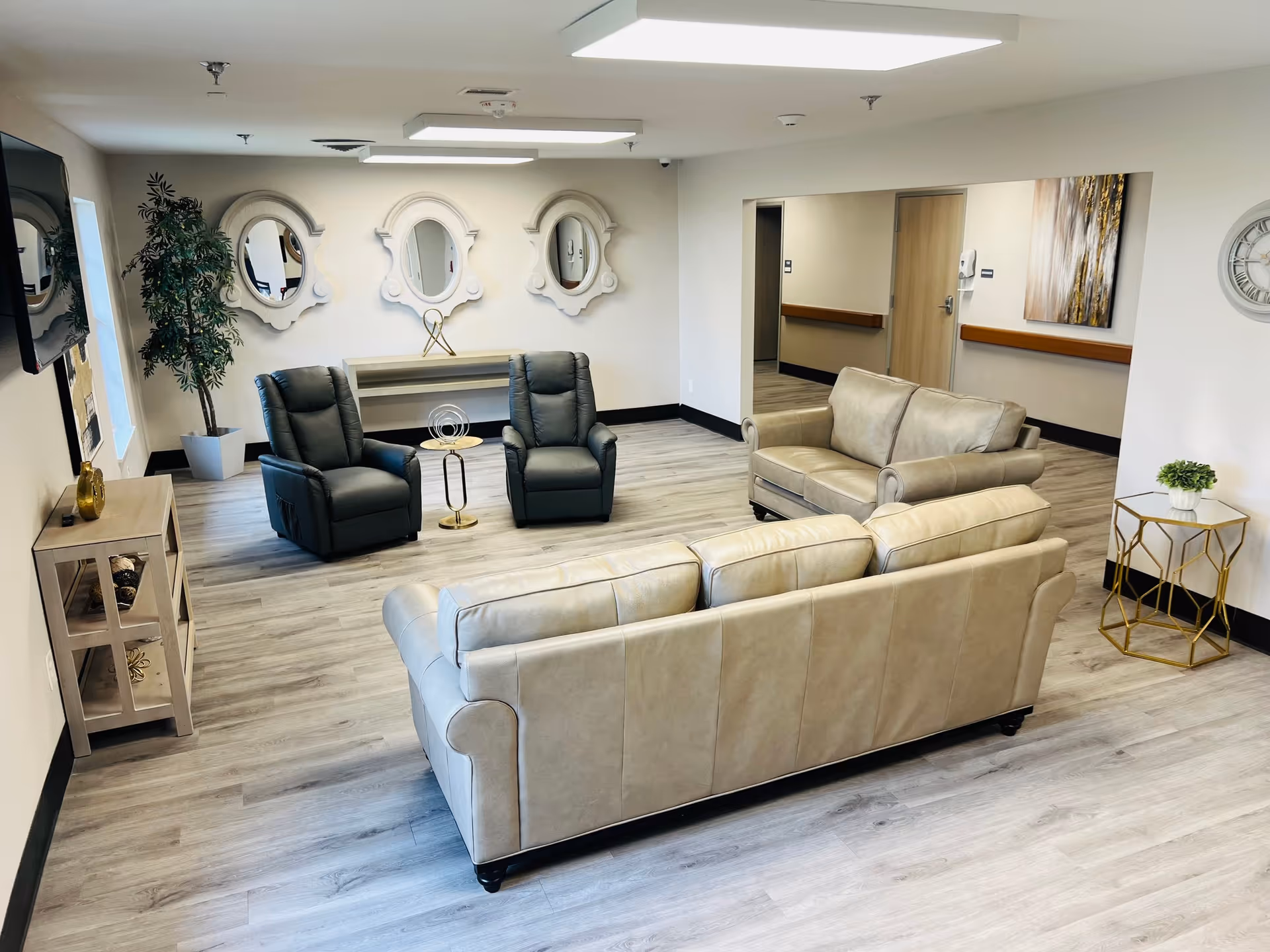 A bright and spacious living room area with light-colored wooden flooring, two beige leather sofas, two dark gray recliner chairs, a small round gold and glass side table, a console table with decorative items, three ornate mirrors on the wall, a potted plant, a wall clock, and a large wall-mounted TV.