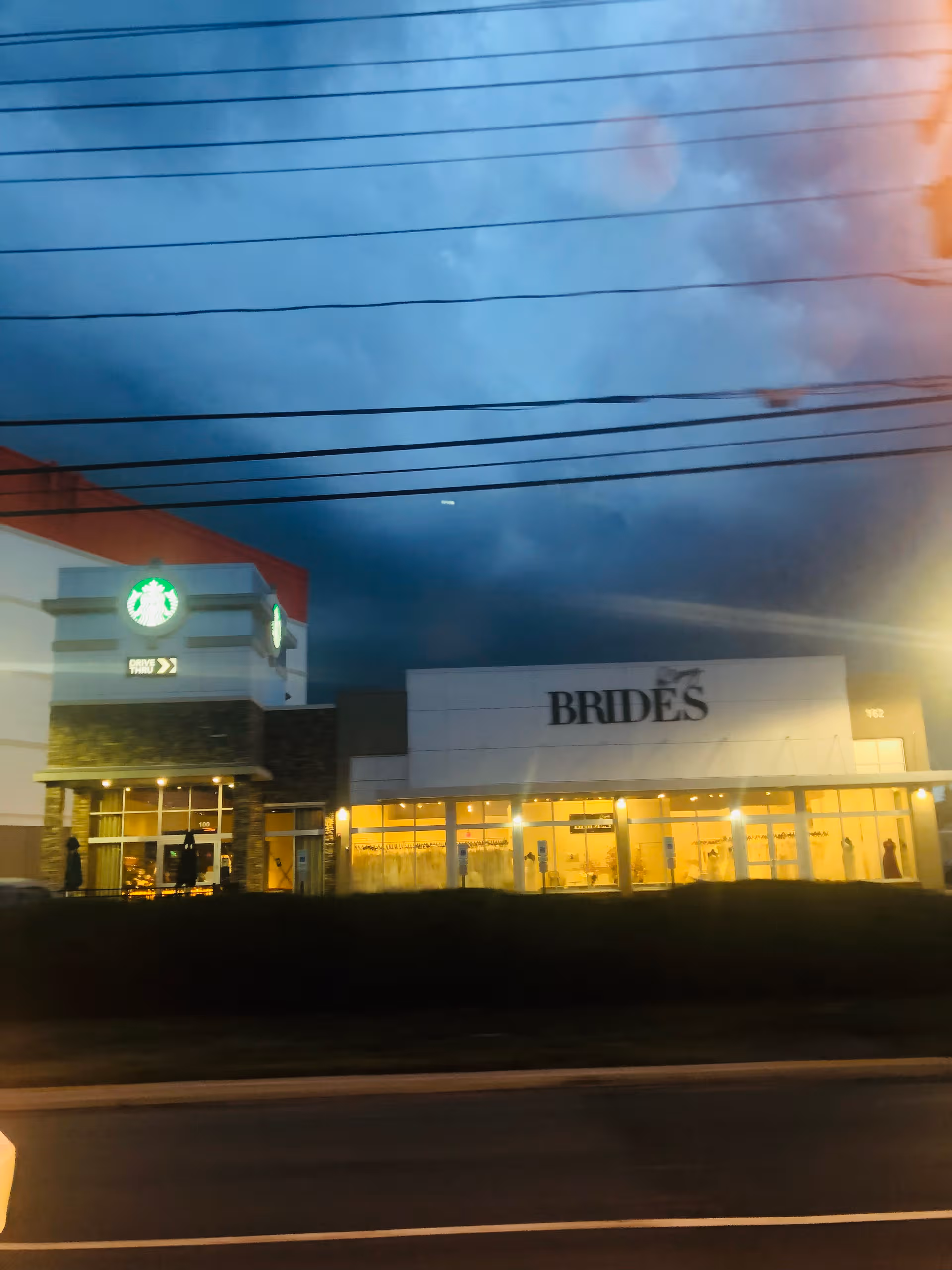 Exterior strip mall frontage at dusk with BRIDES sign and Starbucks storefront.