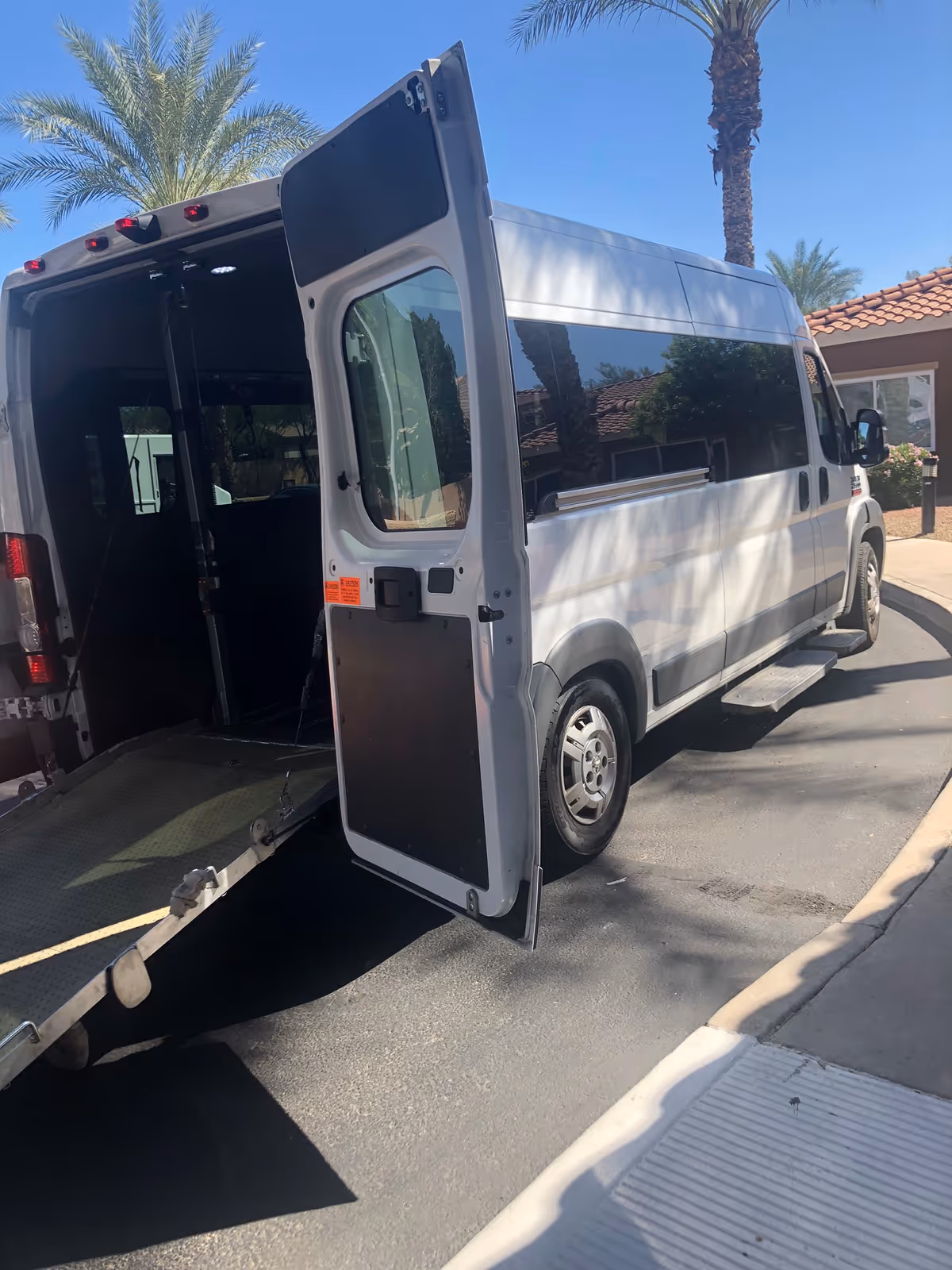 A white wheelchair-accessible van with its rear ramp down, parked outside a facility near palm trees.
