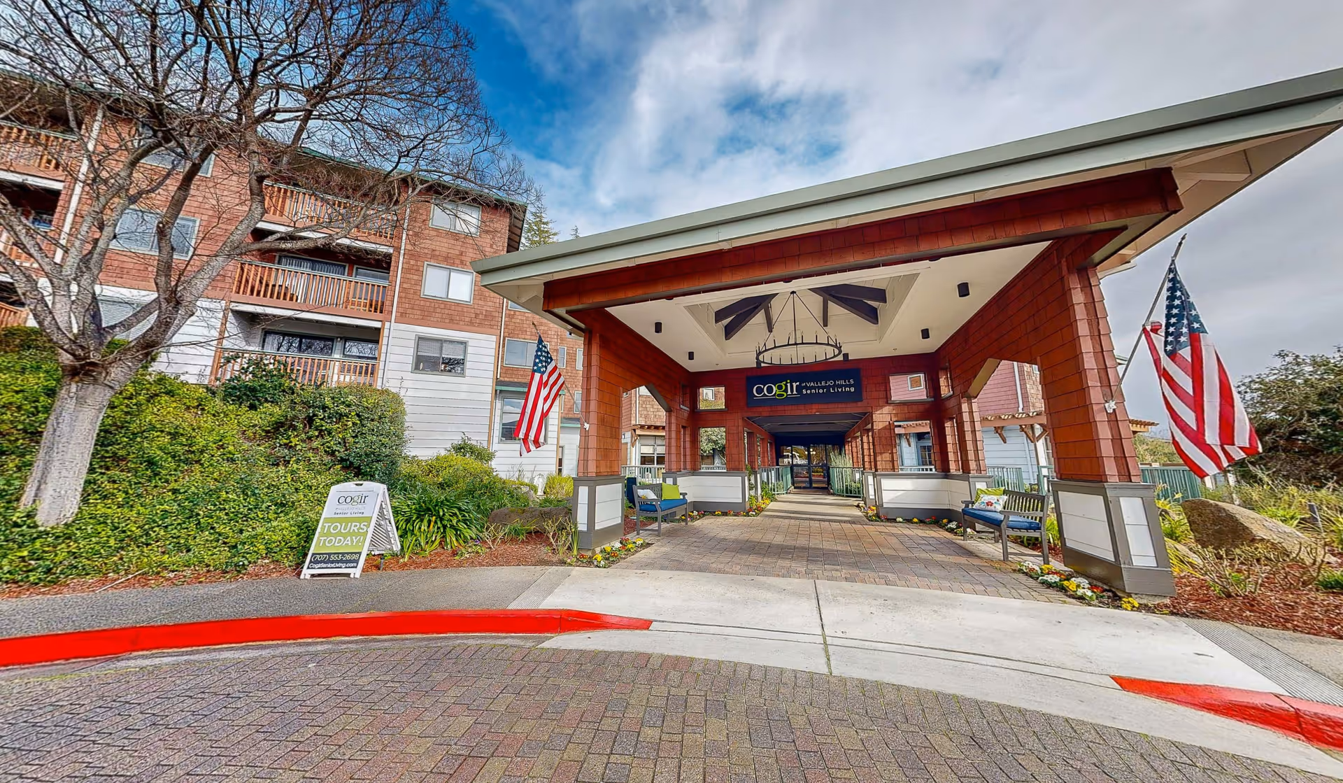 Entrance to Cogir of Vallejo Hills Senior Living facility with a covered walkway, benches, American flags on both sides, and a sign advertising tours. The building is surrounded by greenery and trees under a partly cloudy sky.