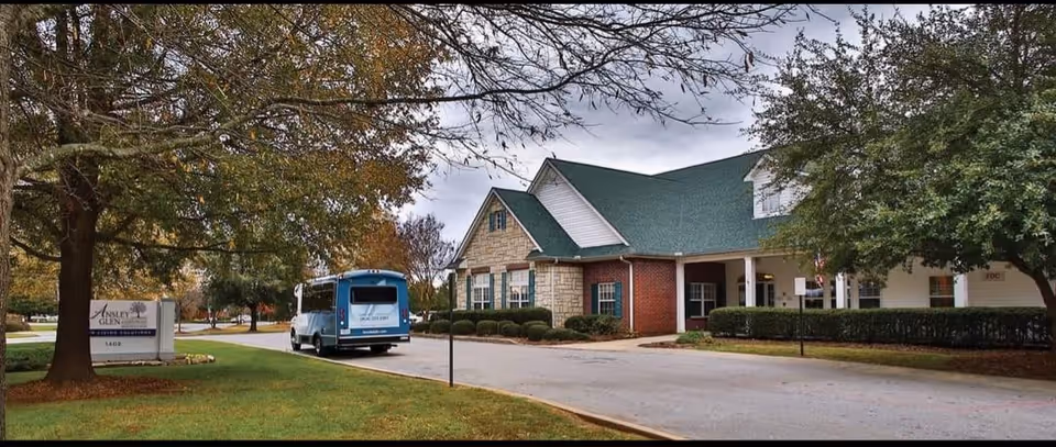 Exterior view of Ansley Glen at Greenwood facility showing a building with a green roof and brick and stone walls, a driveway with a blue shuttle bus parked, and trees with autumn foliage surrounding the area.