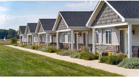 Row of single-story residential units with front porches, white siding, and dark shingled roofs, bordered by a sidewalk and green lawn under a partly cloudy sky.