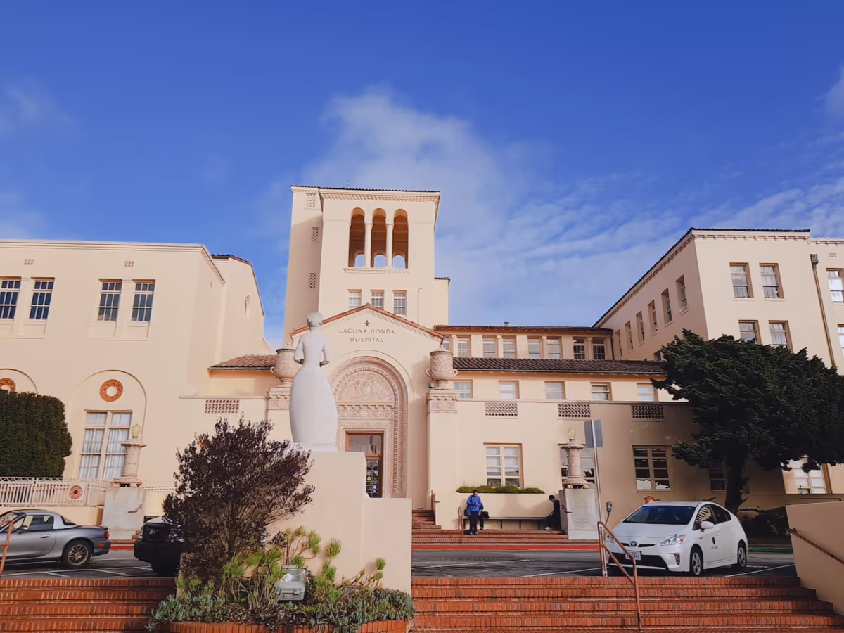Facade of Laguna Honda Hospital with a statue, steps, and parked cars under a blue sky.