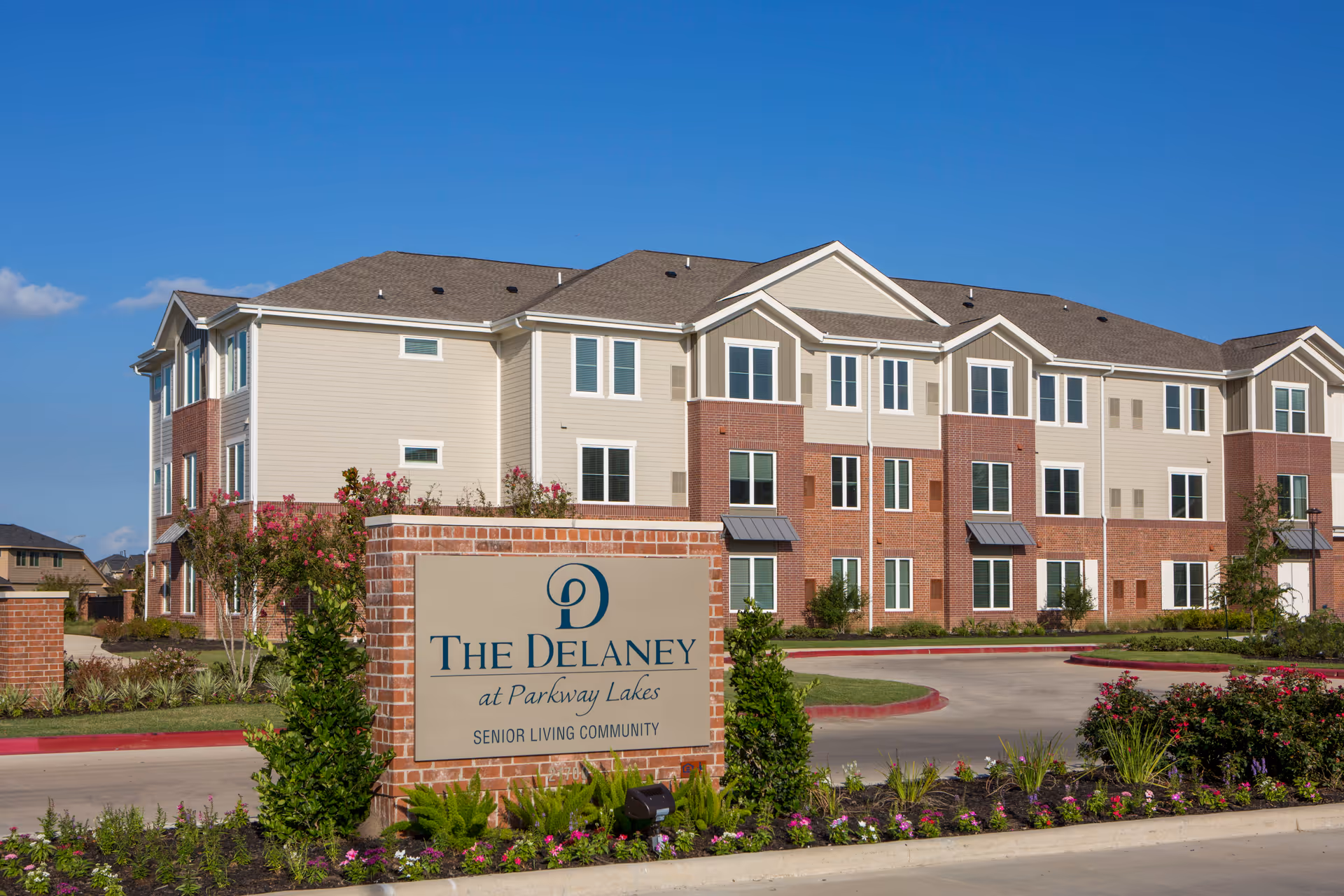 Exterior view of The Delaney at Parkway Lakes senior living community building with a clear blue sky. The building is three stories tall with a combination of beige siding and red brick. In front of the building is a landscaped area with flowers and shrubs, and a large sign displaying the community name.