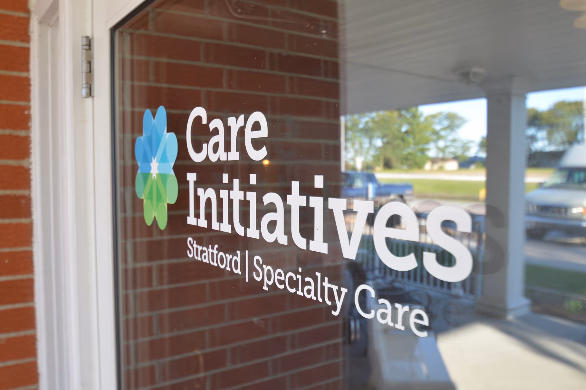 Glass door with the logo and text 'Care Initiatives Stratford Specialty Care' with a brick wall on the left and a porch with vehicles and trees visible through the glass.