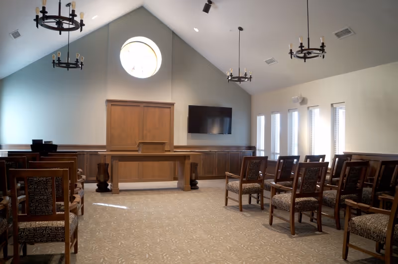 Interior of a small chapel or meeting room with wooden chairs arranged in rows facing a wooden podium and a wall-mounted flat screen TV. The room has a high vaulted ceiling with three black chandeliers and a round window above the podium. Light enters through four narrow windows on the right side.