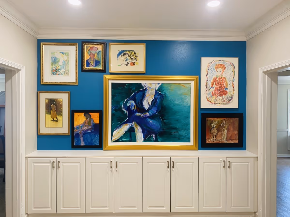 A blue accent wall adorned with eight framed paintings, mostly portraits and abstract figures, above a white cabinet with multiple doors. The wall is flanked by two doorways leading to other rooms with light-colored walls and wooden floors.