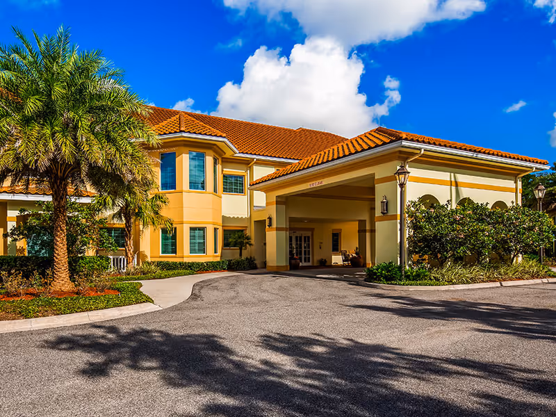 Front entrance of a Mediterranean-style senior living building with a porte-cochère, palm trees, and a bright blue sky.