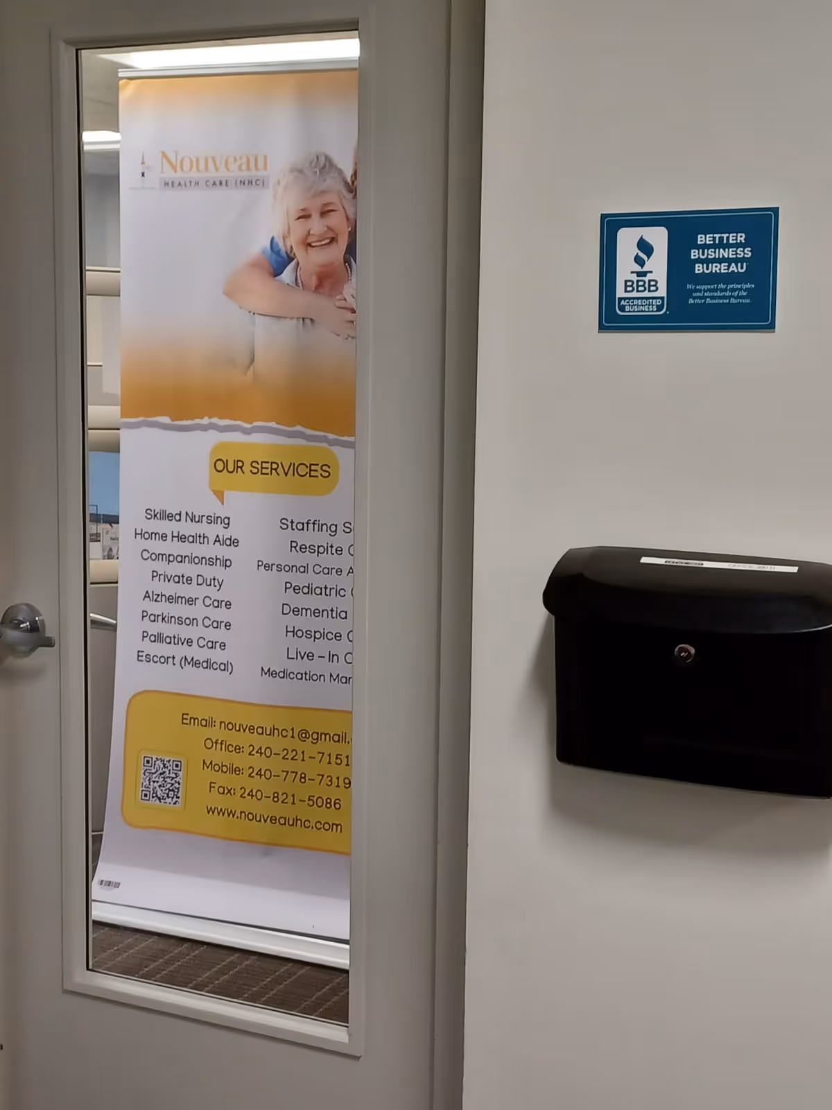 Interior hallway with a door/window displaying a Nouveau Health Care banner next to a BBB sign.