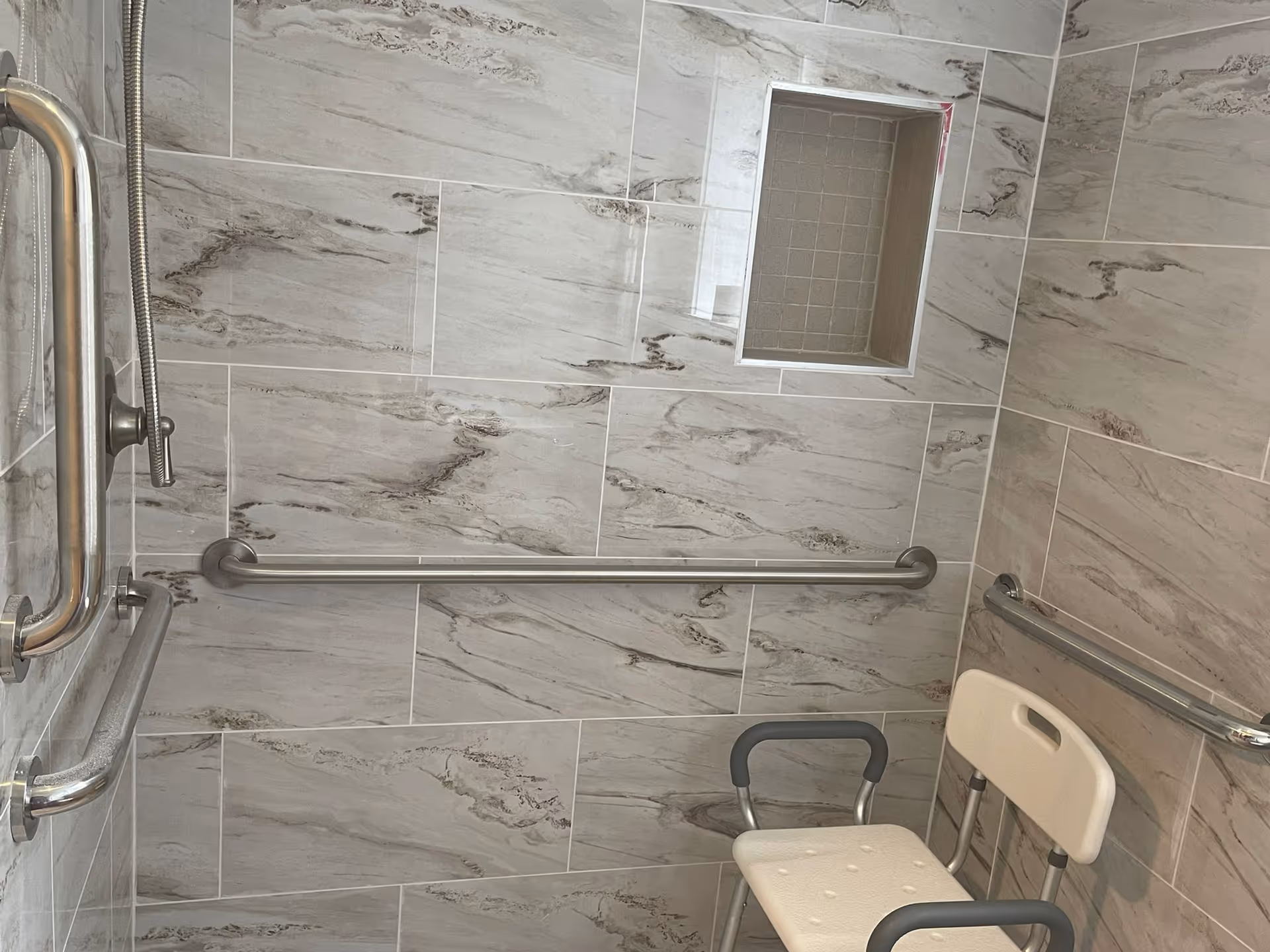 Accessible tiled bathroom with grab bars, a handheld shower, and a small seat.