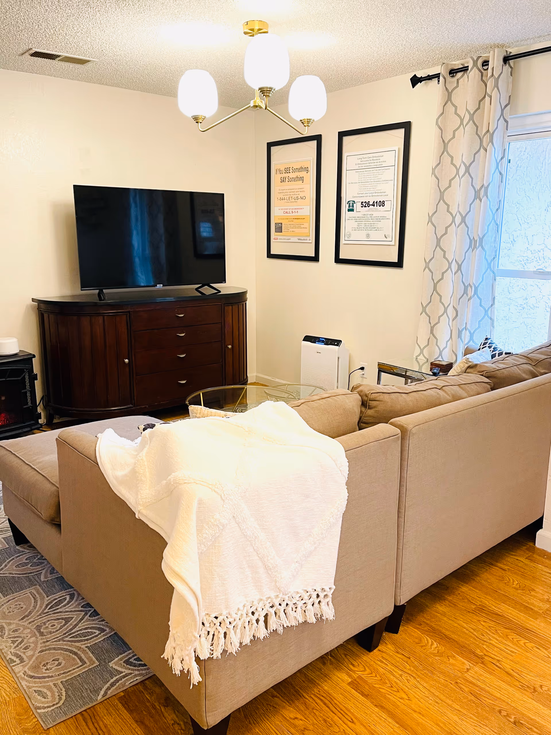 Cozy living room with a beige sectional sofa draped with a white fringed throw blanket, a glass coffee table, a dark wooden TV stand with a flat-screen television, two framed posters on the wall, patterned curtains on the window, and a modern ceiling light fixture.