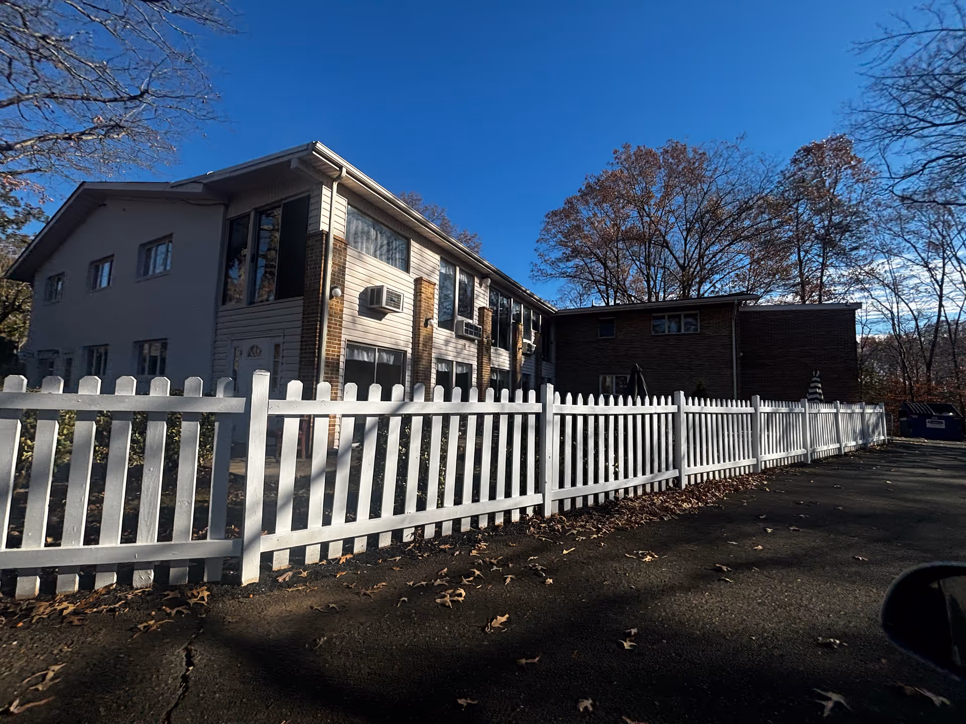 Exterior view of a two-story residential building with white siding and brick accents, surrounded by a white picket fence. The scene includes leafless trees and a clear blue sky.