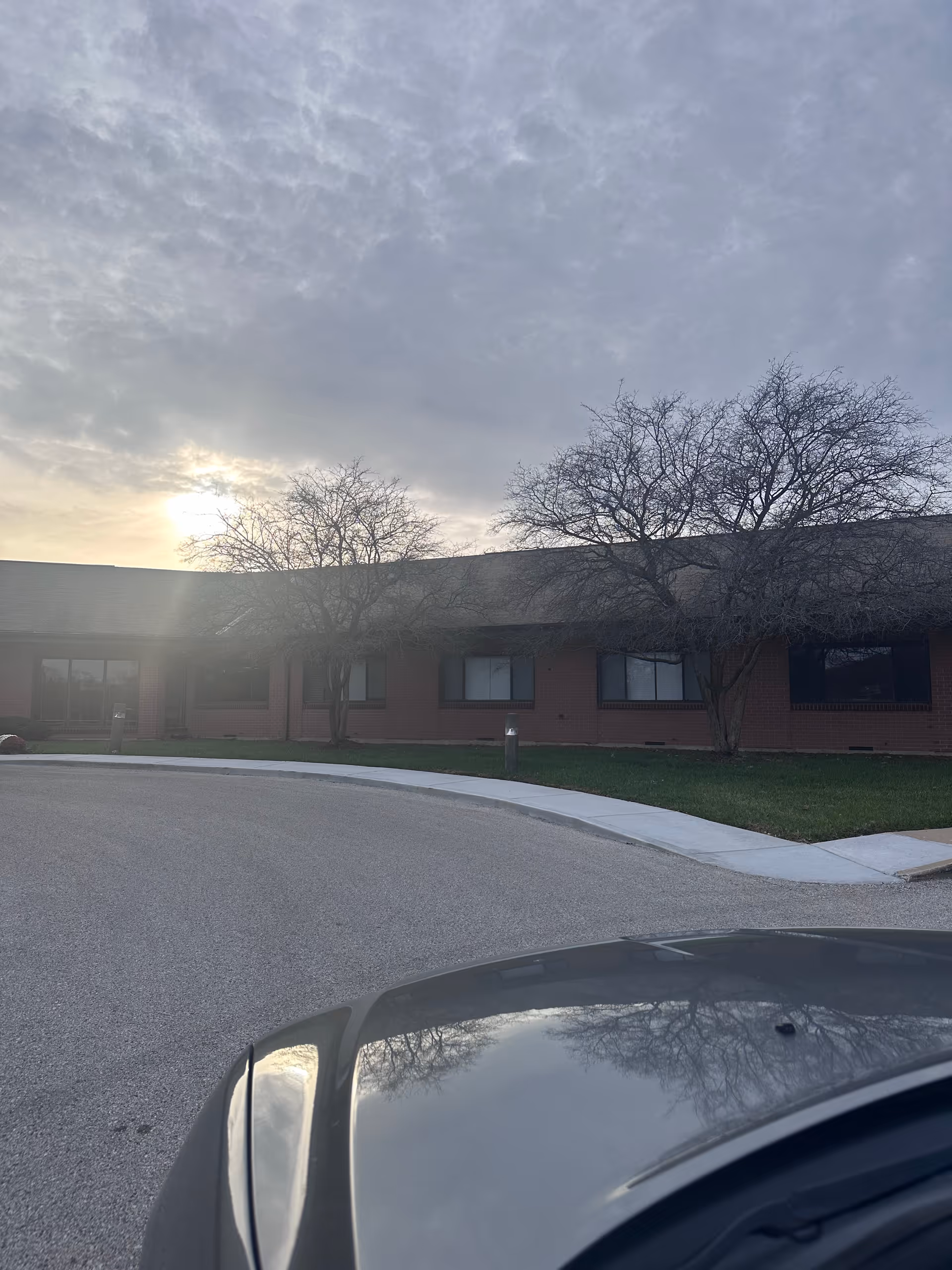 Exterior view of a single-story brick building with several windows and leafless trees in front. The sky is cloudy with the sun partially visible. A curved driveway and a portion of a car's hood are visible in the foreground.